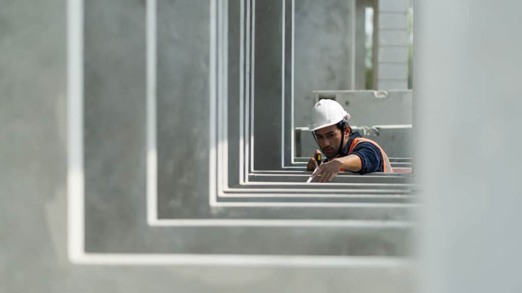 Construction worker in a hard hat and safety vest inspecting precast concrete structures, framed through a series of rectangular concrete openings.